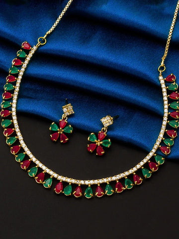 Gold-tone necklace and matching earrings set with alternating red and green teardrop gemstones and clear stones, displayed on a dark surface with a blue satin cloth.