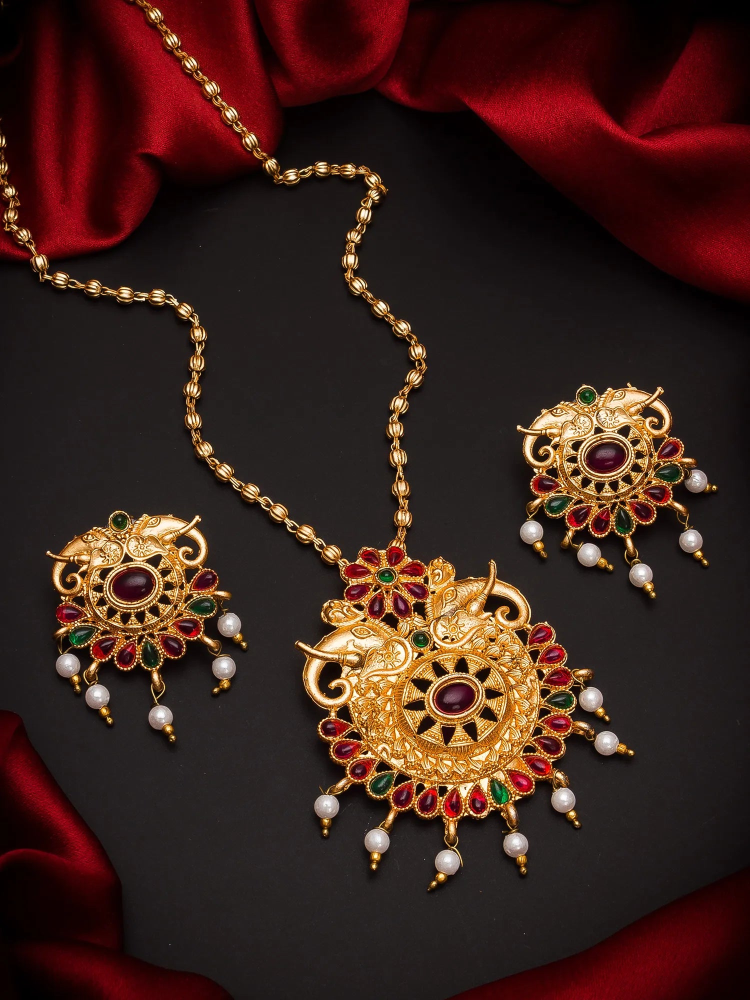 Gold-toned traditional jewelry set featuring a necklace, earrings, and a pendant with ornate filigree, red and green stone accents, and dangling white pearl beads, arranged on a dark surface with red satin fabric.