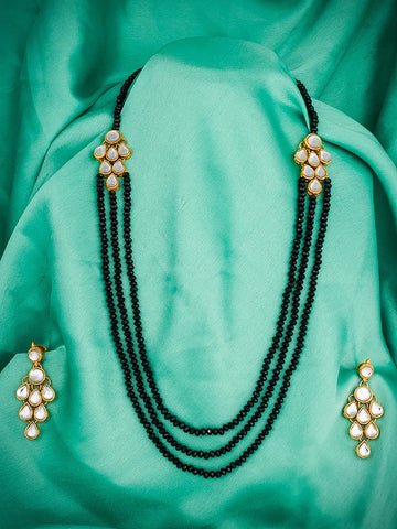 A black beaded necklace with gold-toned and white stone embellishments, displayed with matching earrings on a draped mint green satin fabric background.