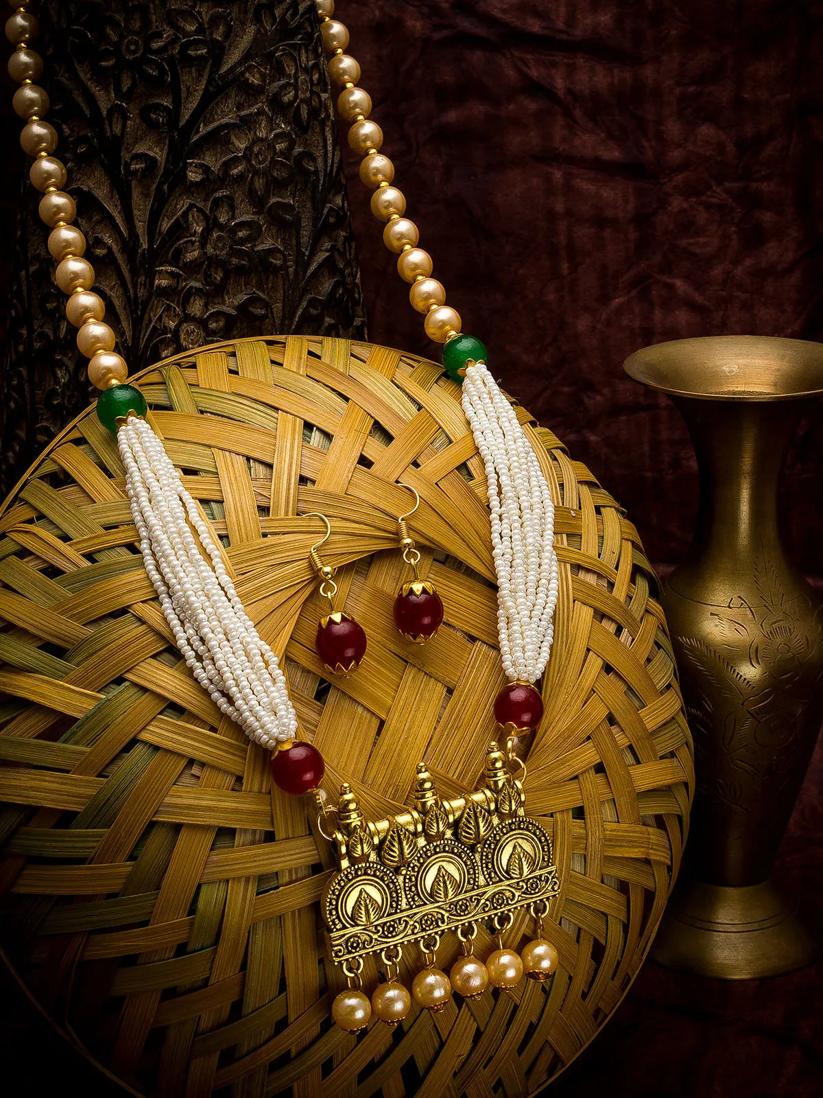 A traditional Indian jewelry set featuring a necklace and matching earrings, displayed on a woven bamboo basket. The necklace has white beaded tassels, red marigold beads, and a gold-toned pendant with hanging pearl-like drops.