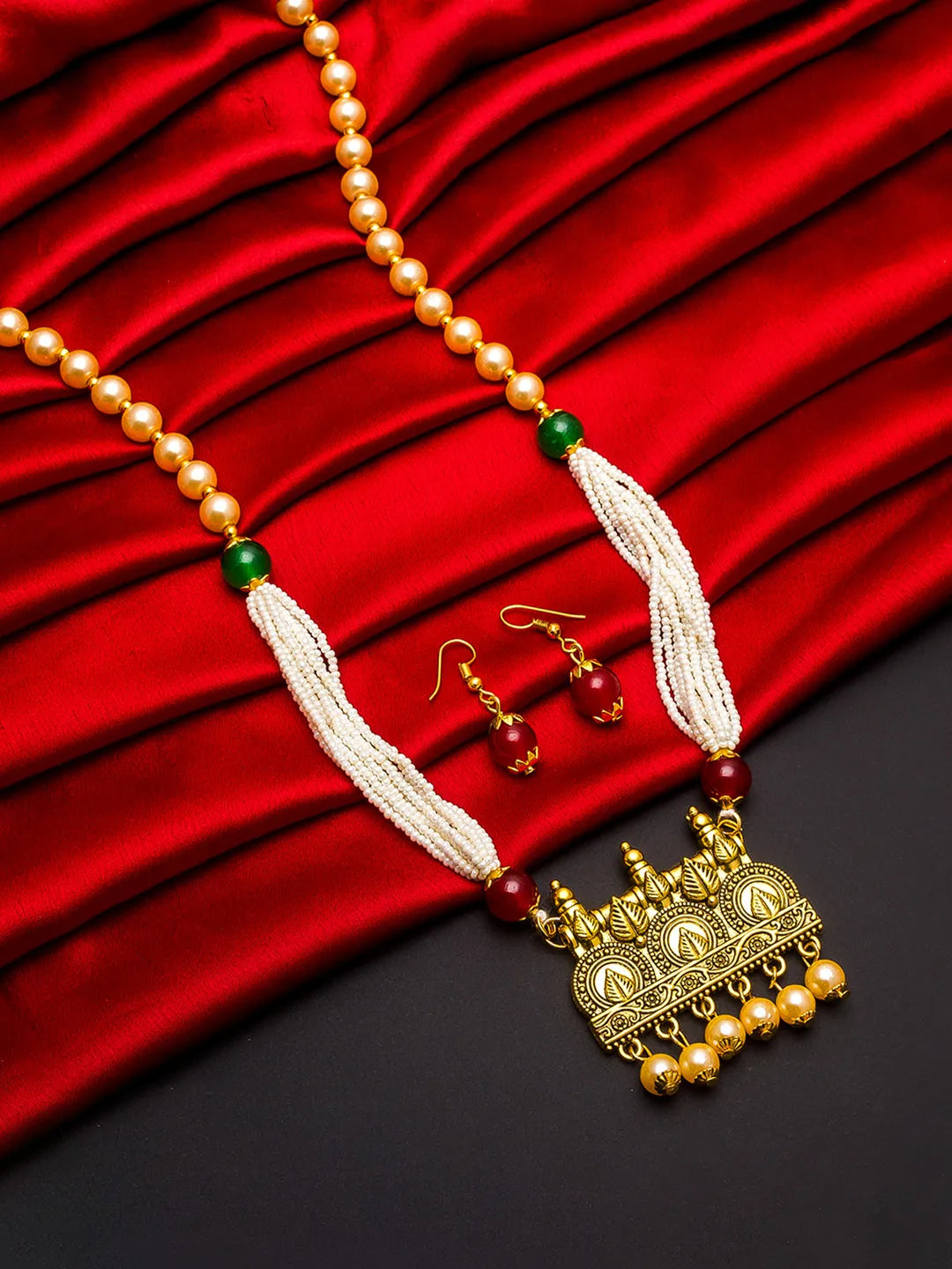 A traditional Indian jewelry set featuring a gold-plated pendant necklace with white pearl strands, red and green beads, and matching gold earrings, displayed on a draped red satin fabric background.