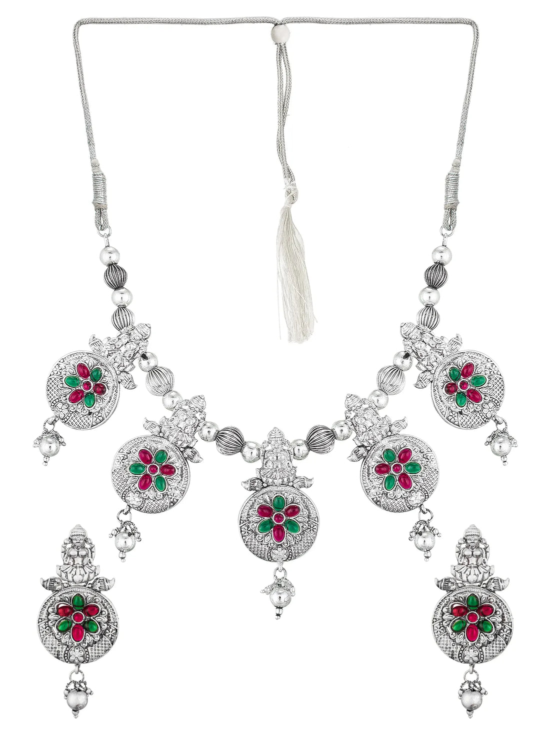 An ornate silver-toned jewelry set displayed on a black mannequin bust, including a necklace, matching earrings, and a pair of dangling earrings, decorated with silver beads, floral motifs, and red and green stone accents. A vase of flowers is visible on the left against a teal background.
