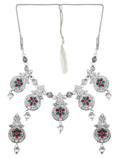 Silver-toned traditional Indian jewelry set featuring a necklace and matching earrings with ornate filigree work and floral motifs in red and green stones.