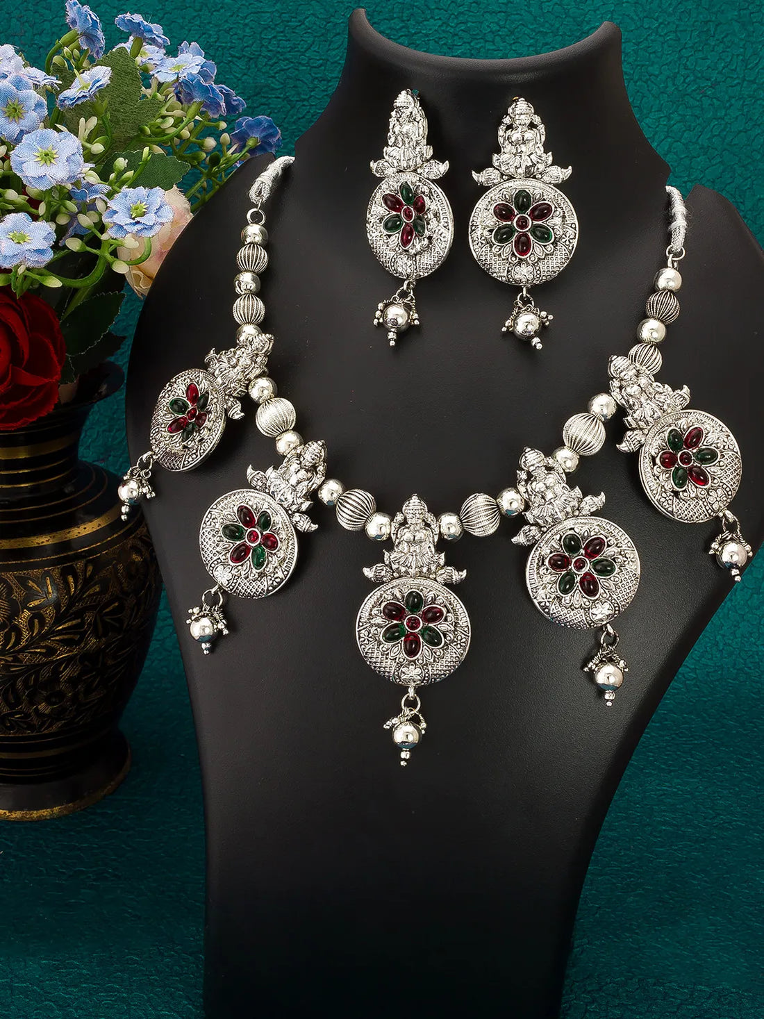 An ornate silver-toned jewelry set displayed on a black mannequin bust, including a necklace, matching earrings, and a pair of dangling earrings, decorated with silver beads, floral motifs, and red and green stone accents. A vase of flowers is visible on the left against a teal background.