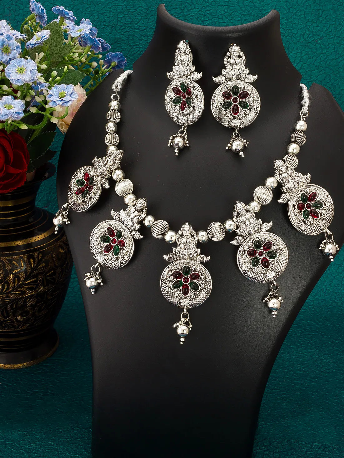 An ornate silver-toned jewelry set displayed on a black mannequin bust, including a necklace, matching earrings, and a pair of dangling earrings, decorated with silver beads, floral motifs, and red and green stone accents. A vase of flowers is visible on the left against a teal background.