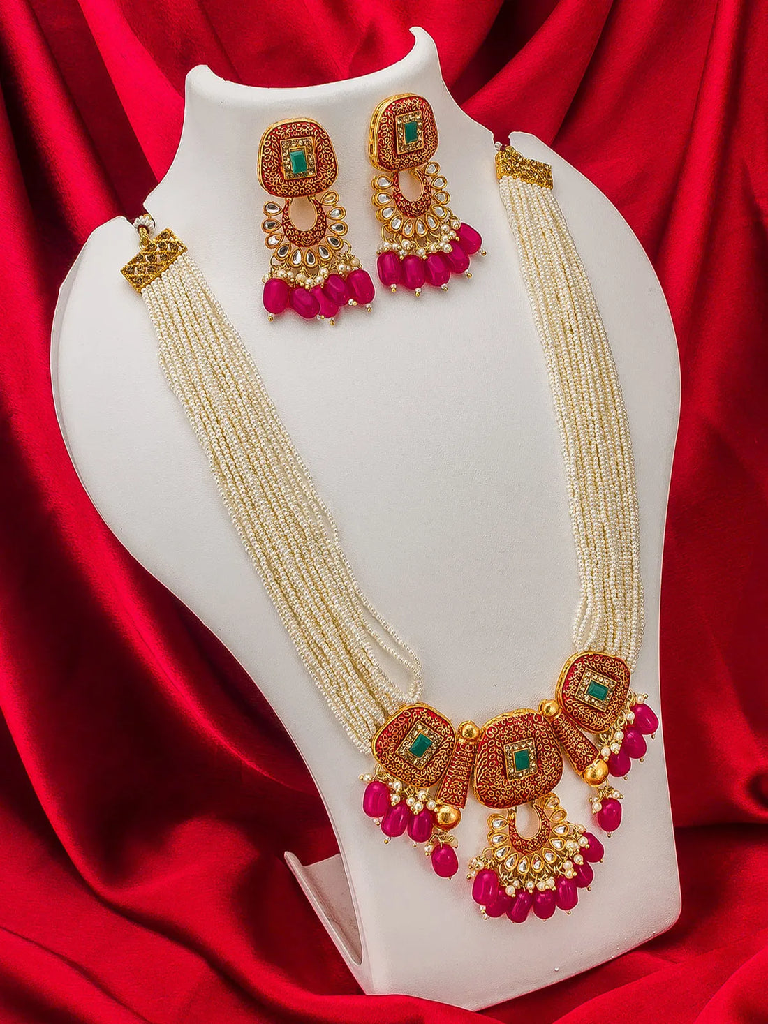 A traditional Indian jewelry set featuring a multi-strand cream bead necklace and matching earrings with gold-plated details, green stones, and bright pink bead drops, displayed on a white jewelry bust against a red satin background.