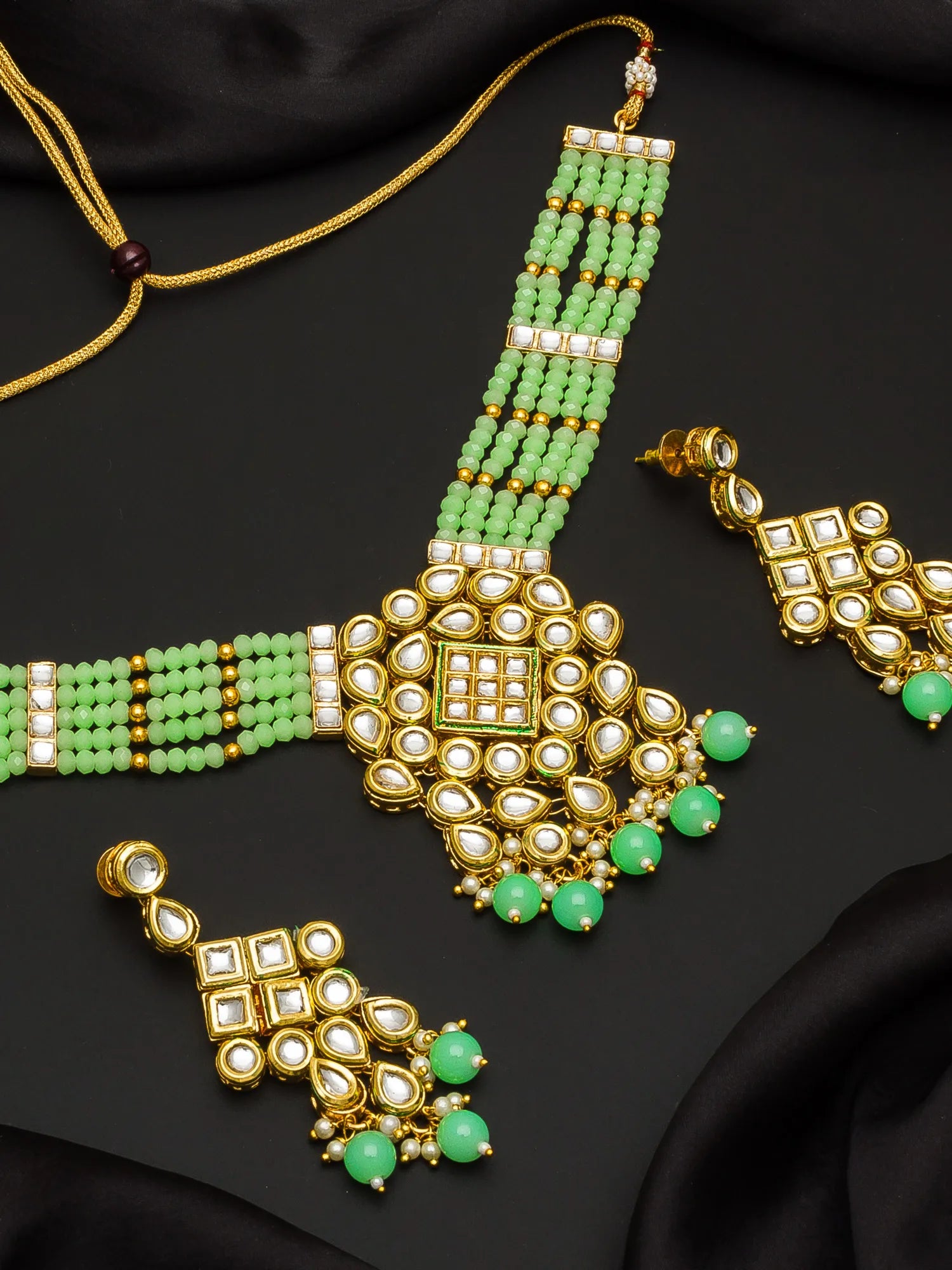 Gold-plated Indian bridal jewelry set featuring a necklace, matching earrings, and a bracelet, embellished with light green beads, white stones, and small gold accents, displayed on a black satin fabric background.