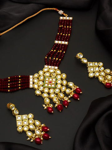 Gold-plated Indian traditional jewelry set featuring a multi-strand beaded necklace, a large matching pendant, and two matching earrings, decorated with white stones, red beads, and a small green accent, displayed on a black satin background.