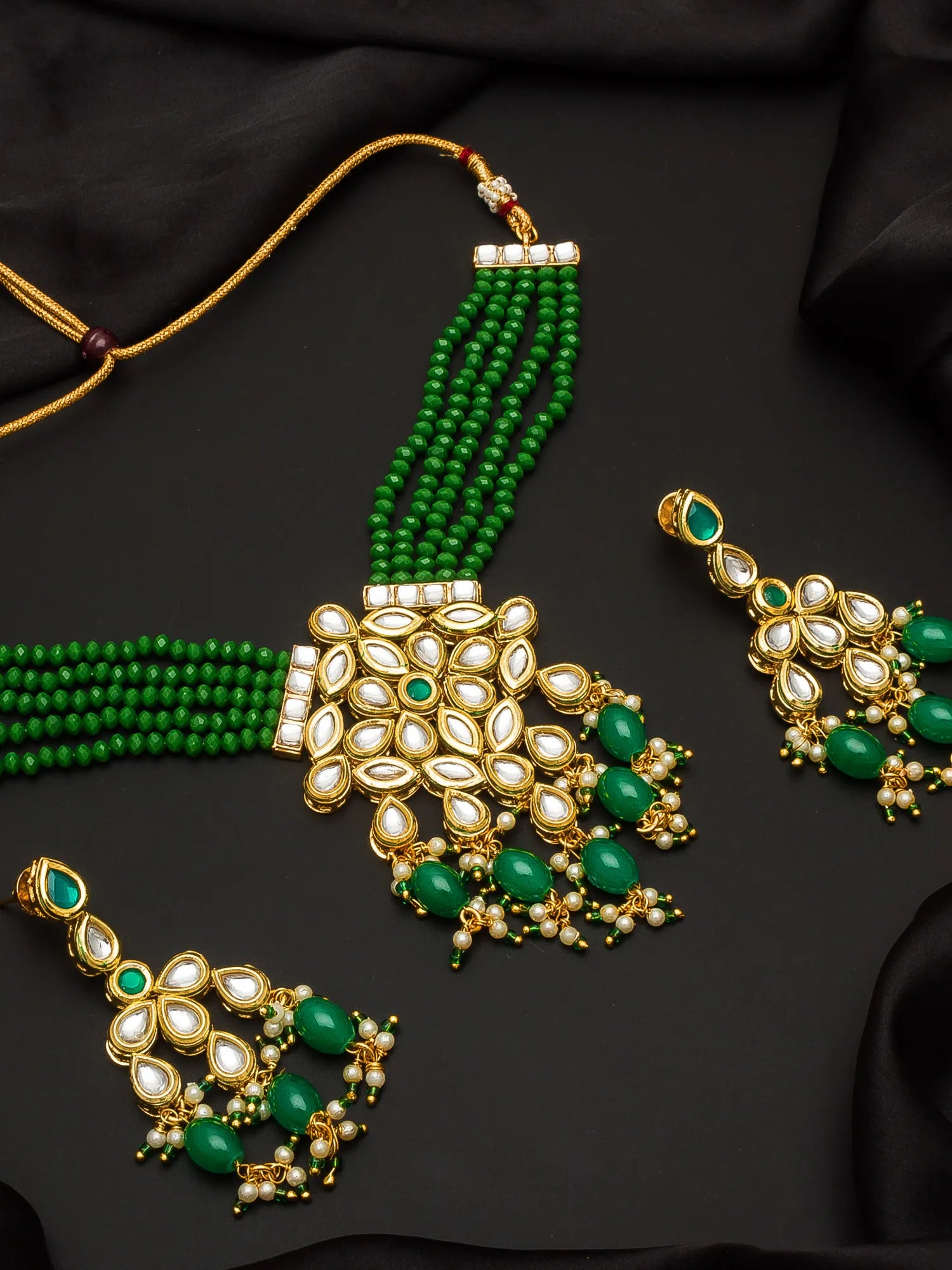 Gold and green traditional Indian jewelry set featuring a multi-strand beaded necklace, a large central pendant, and two matching earrings, arranged on a black fabric background.
