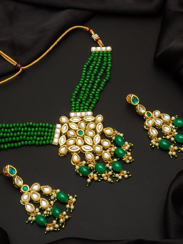 Gold and green traditional Indian jewelry set featuring a multi-strand beaded necklace, a large central pendant, and two matching earrings, arranged on a black fabric background.