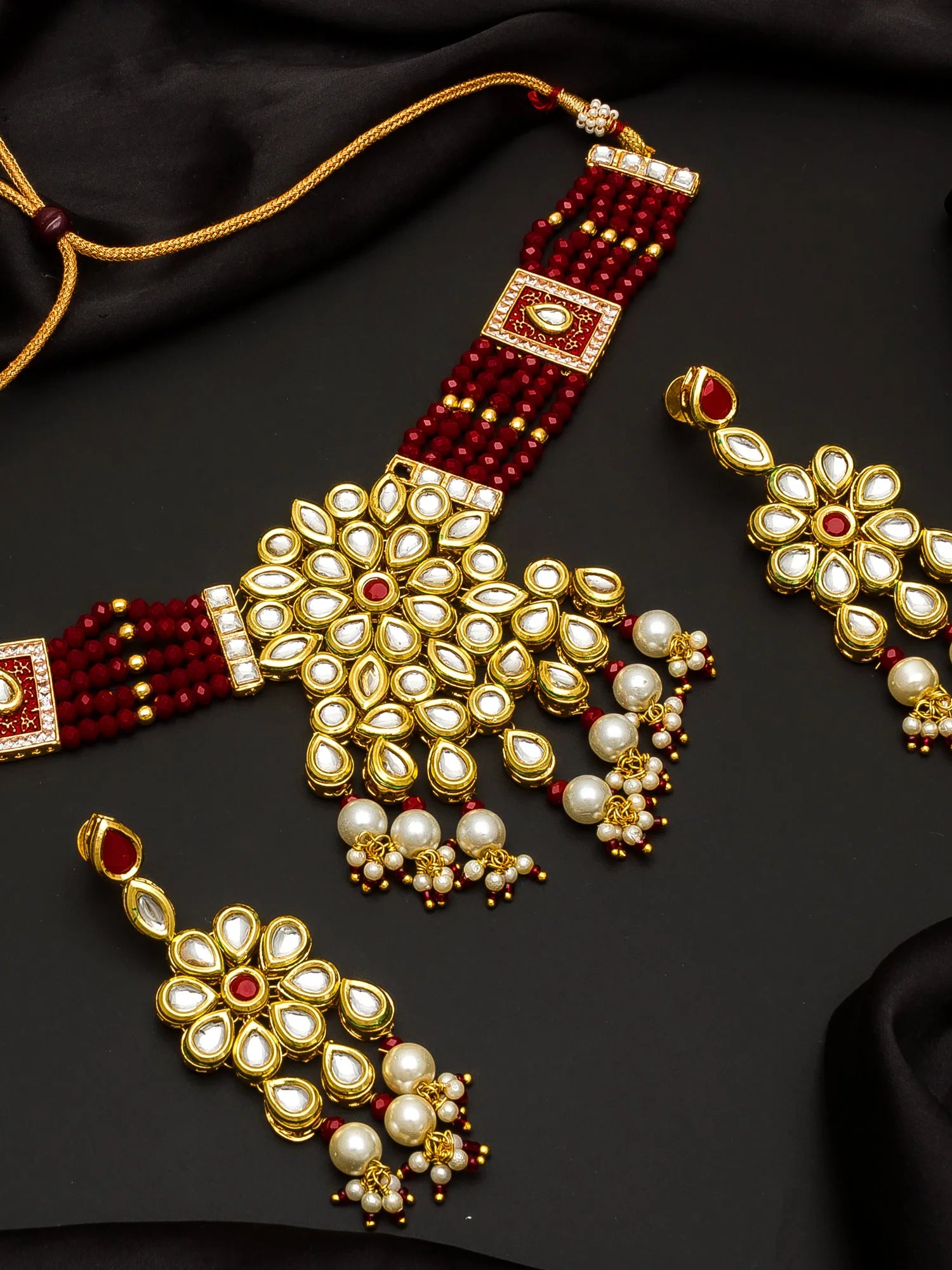 Gold and red beaded traditional Indian jewelry set featuring a necklace, earrings, and a maang tikka, embellished with white faux pearls and set against a black satin background.