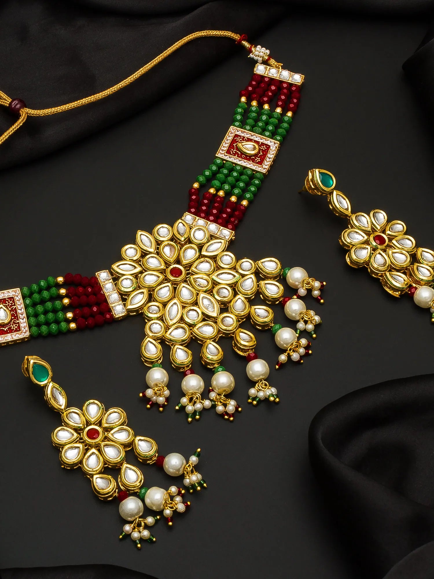 An ornate Indian jewelry set featuring a gold-plated necklace and matching earrings decorated with white faux pearls, green and red beads, and small red accent stones, displayed on a black satin background.