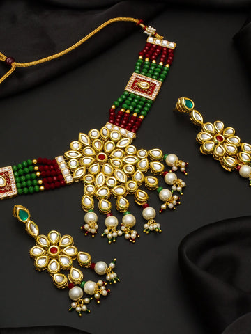 An ornate Indian jewelry set featuring a gold-plated necklace and matching earrings decorated with white faux pearls, green and red beads, and small red accent stones, displayed on a black satin background.