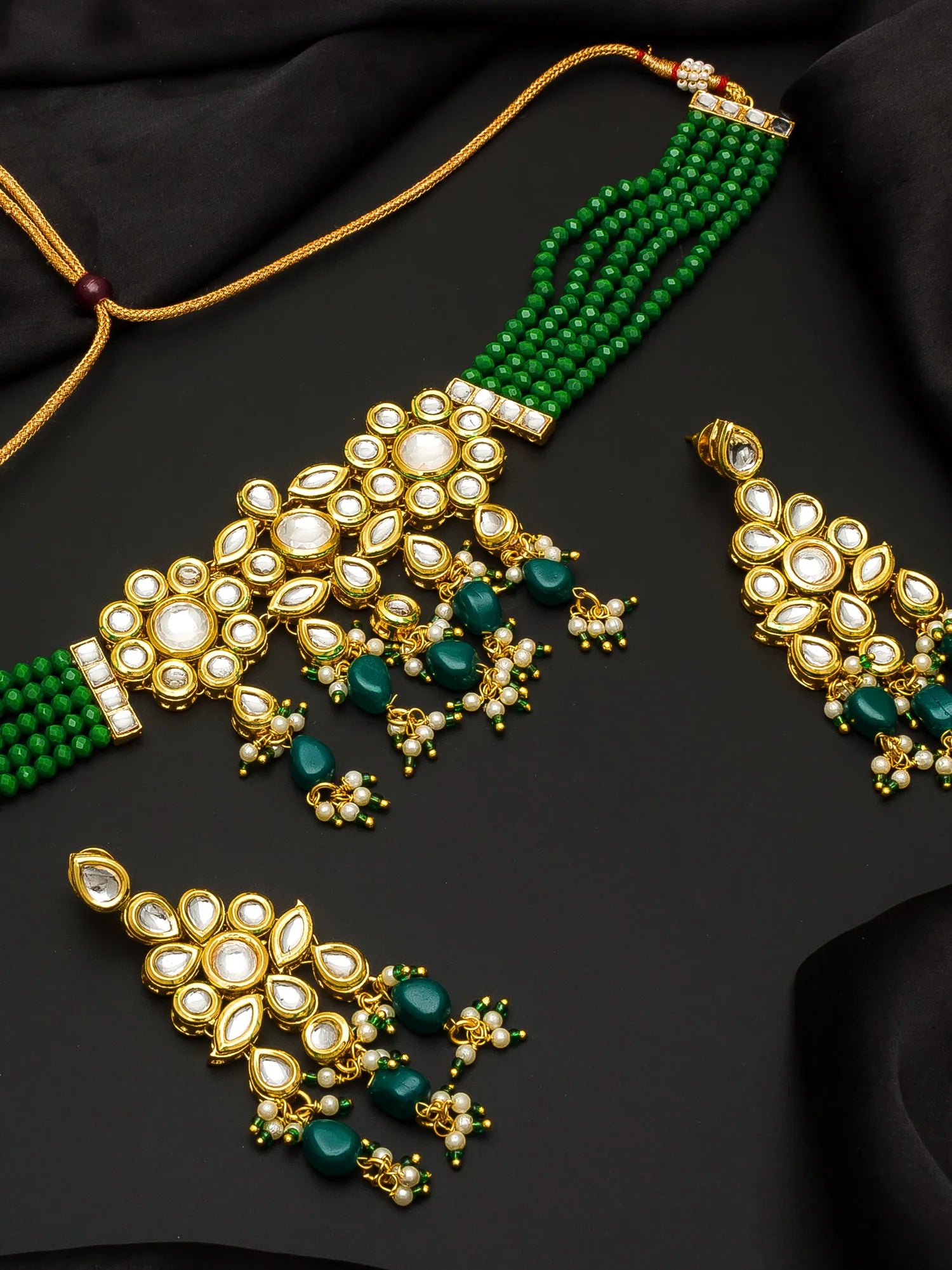 Gold and green traditional Indian jewelry set featuring a necklace, earrings, and a choker with white stones and green beads, arranged on a black fabric background.