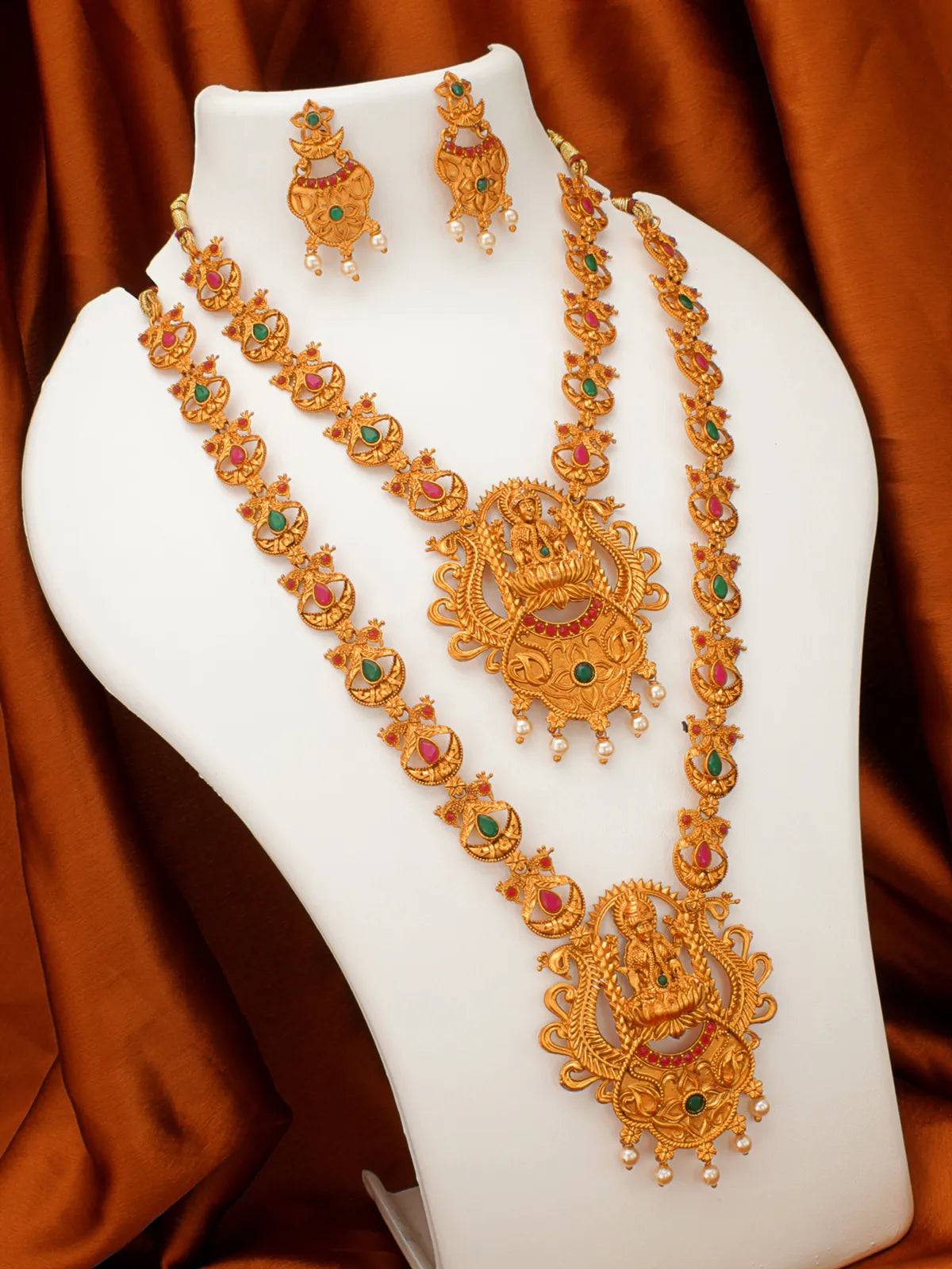 An ornate gold-toned traditional jewelry set displayed on a white mannequin bust, featuring a layered necklace with a large central pendant, matching earrings, and small pearl-like accents, set against a draped rust-brown fabric background.