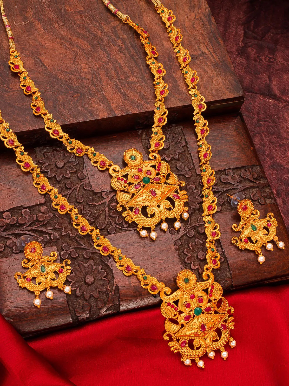 An ornate gold jewelry set featuring a necklace, pendant, earrings, and matching bracelet, decorated with red and green stones and small pearl-like drops, displayed on a carved dark wooden box over a red fabric background.