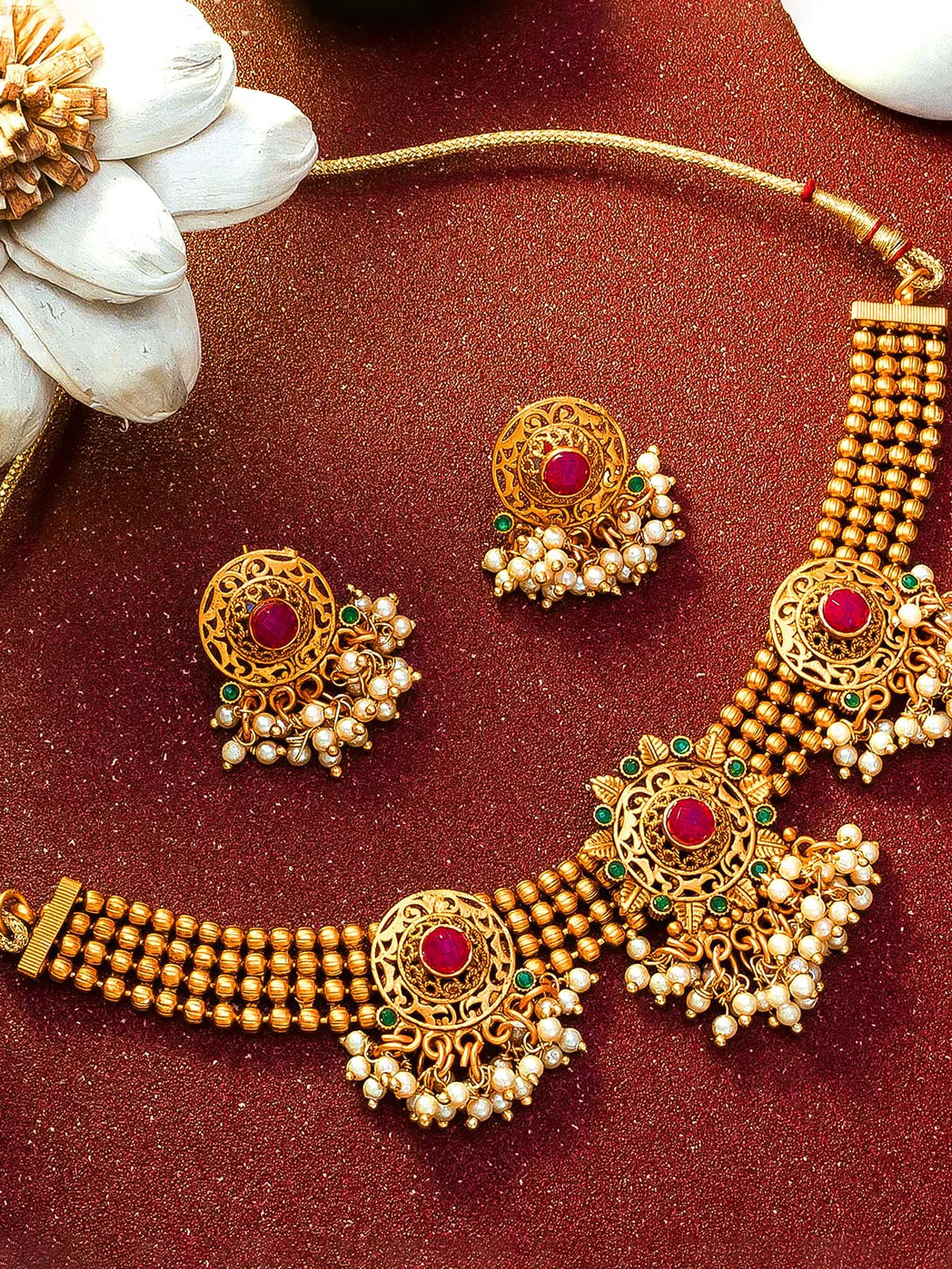 Gold-toned traditional jewelry set with a necklace and matching earrings, featuring red gemstone centers, green stone accents, and white pearl-like drops, arranged on a textured dark red surface with decorative white flower props.