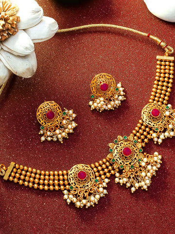 Gold-toned traditional jewelry set with a necklace and matching earrings, featuring red gemstone centers, green stone accents, and white pearl-like drops, arranged on a textured dark red surface with decorative white flower props.
