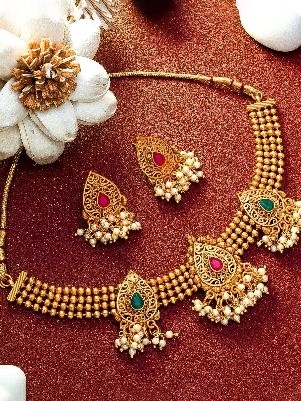 Gold-toned traditional Indian jewelry set featuring a multi-strand beaded necklace and matching earrings with red and green stone accents and pearl-like dangling beads, arranged on a textured dark red surface with decorative white flowers.