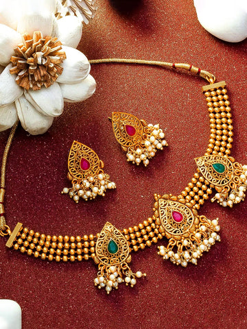 Gold-toned traditional Indian jewelry set featuring a multi-strand beaded necklace and matching earrings with red and green stone accents and pearl-like dangling beads, arranged on a textured dark red surface with decorative white flowers.