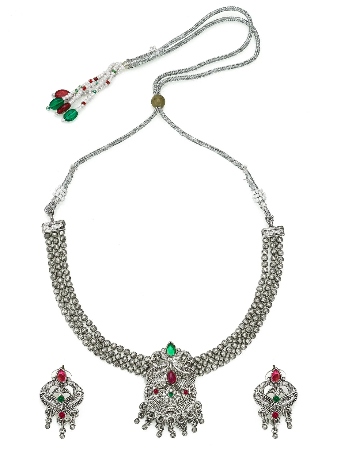 A silver-toned traditional Indian jewelry set featuring a beaded necklace, matching earrings, and a large pendant, decorated with red and green stones, displayed on a draped red fabric background.