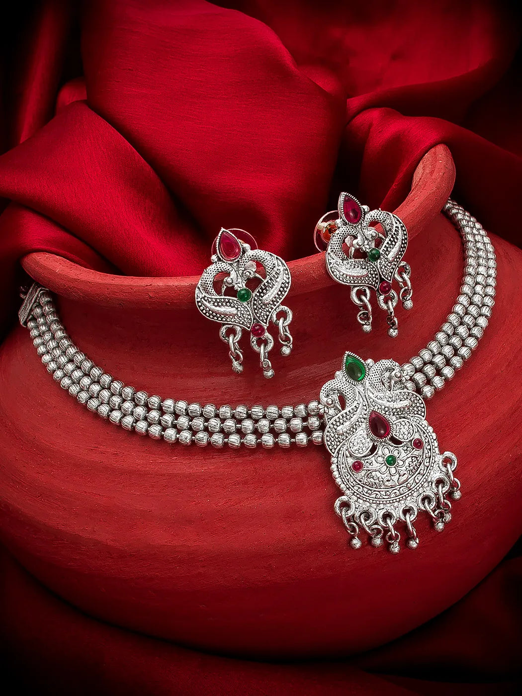 A silver-toned traditional Indian jewelry set featuring a beaded necklace, matching earrings, and a large pendant, decorated with red and green stones, displayed on a draped red fabric background.