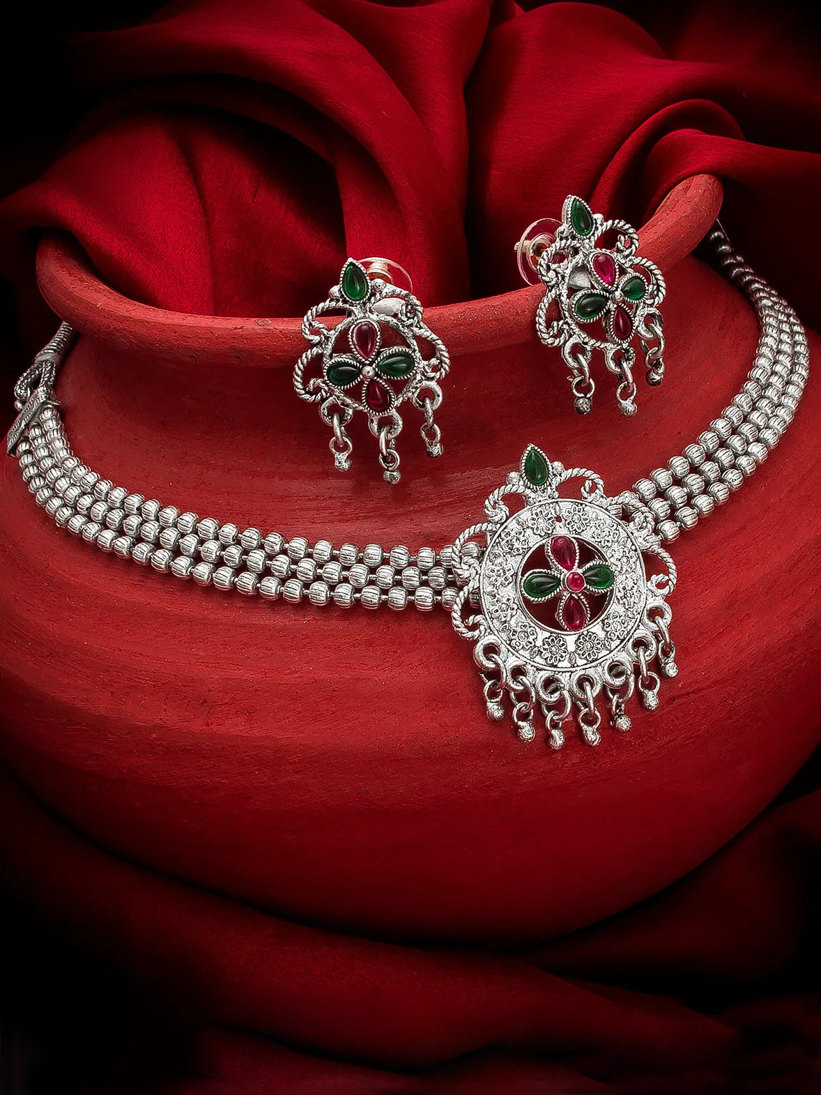 Silver jewelry set with red and green stones on a red fabric background
