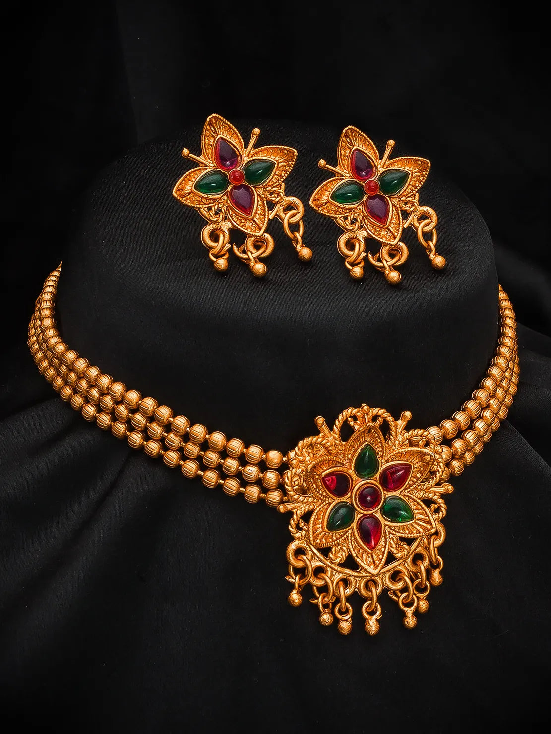 Gold-toned traditional jewelry set featuring a beaded necklace with an ornate central pendant and matching butterfly-shaped earrings, decorated with red and green stones, displayed on a black fabric stand.