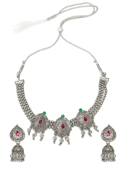A traditional Indian silver-toned jewelry set featuring a necklace and matching earrings. The necklace has a textured silver chain with a central ornate silver pendant decorated with three red teardrop gemstones and small green stone accents.