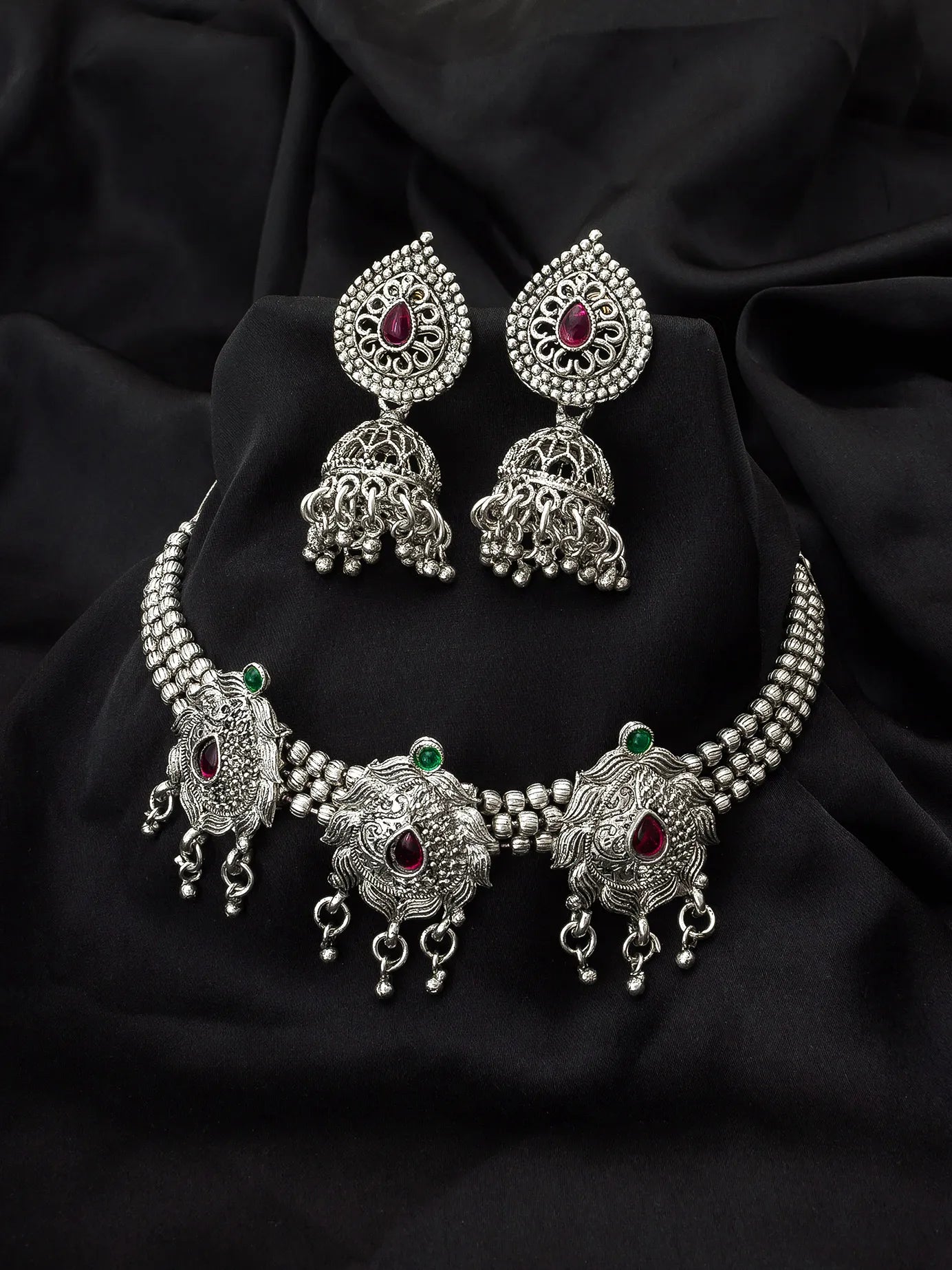Silver-toned traditional Indian jewelry set featuring a choker necklace, matching earrings, and a waist chain, embellished with silver filigree, beaded borders, and red and green stone accents, displayed on a draped black fabric background.