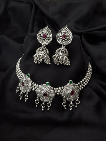 Silver-toned traditional Indian jewelry set featuring a choker necklace, matching earrings, and a waist chain, embellished with silver filigree, beaded borders, and red and green stone accents, displayed on a draped black fabric background.