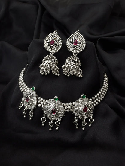 Silver-toned traditional Indian jewelry set featuring a choker necklace, matching earrings, and a waist chain, embellished with silver filigree, beaded borders, and red and green stone accents, displayed on a draped black fabric background.