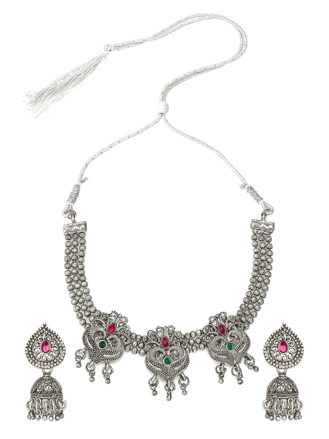 Silver-toned Indian jewelry set featuring a choker necklace, matching earrings, and a headpiece, decorated with intricate filigree work and small red and green gemstones, displayed on a black fabric background.