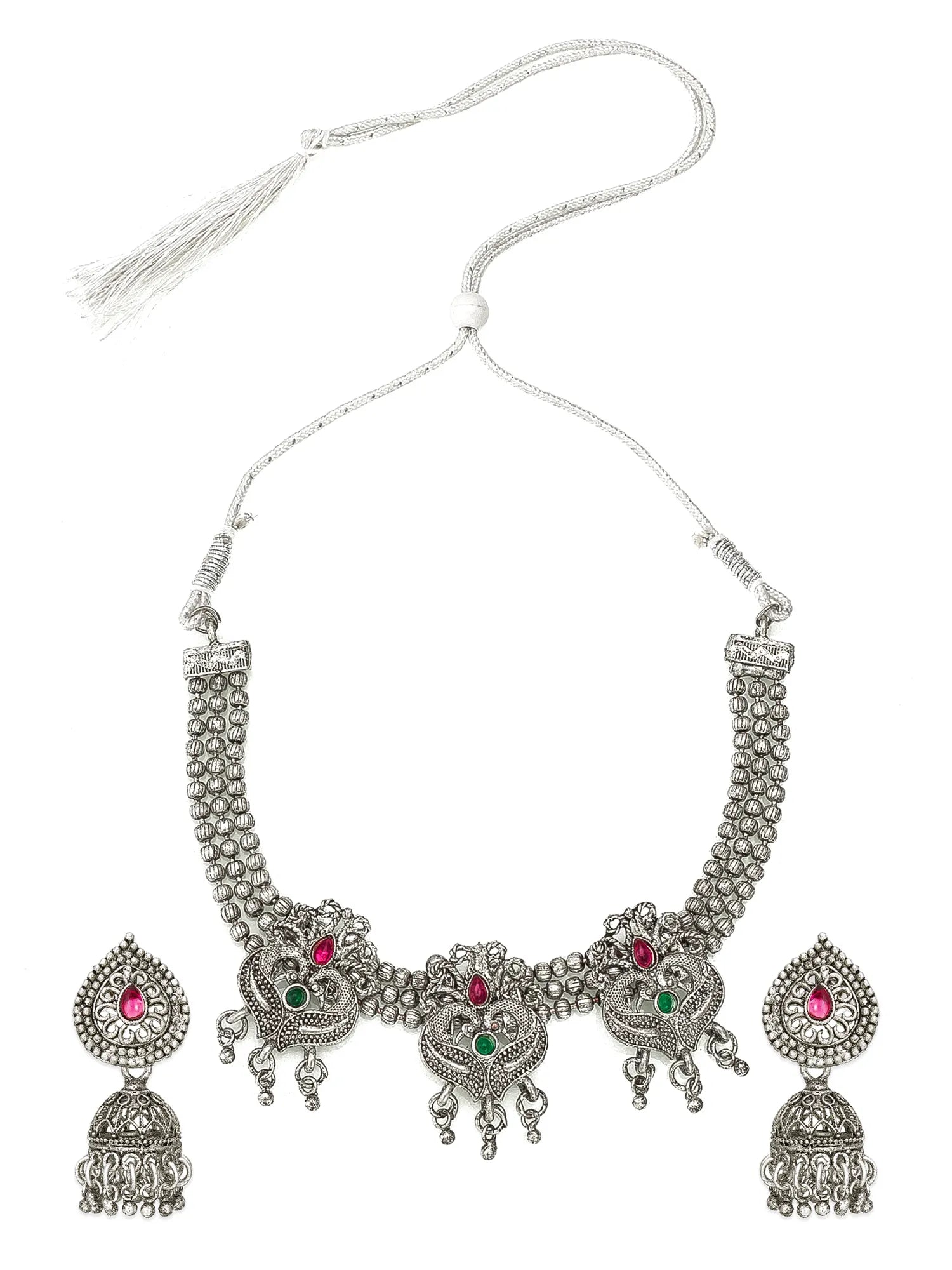 An ornate silver-toned traditional jewelry set featuring a necklace with a tassel and matching drop earrings, decorated with silver filigree, red and green stone accents, and dangling jhumka-style charms, shown on a plain white background.