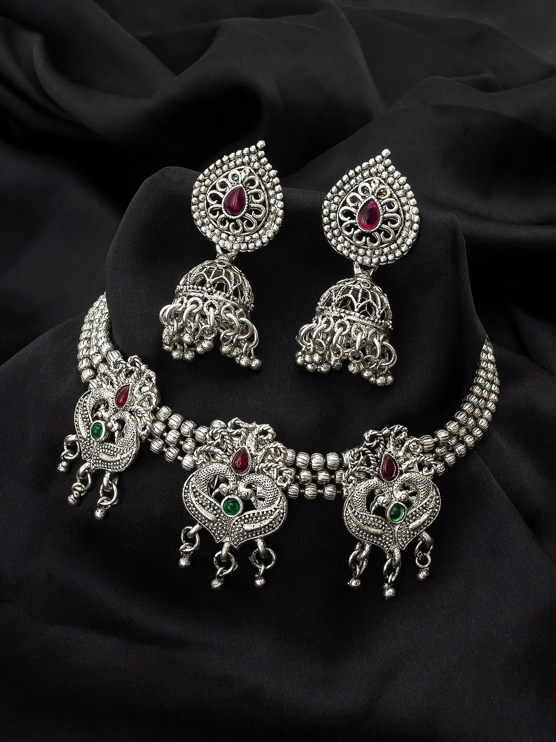 Silver-toned Indian jewelry set featuring a choker necklace, matching earrings, and a headpiece, decorated with intricate filigree work and small red and green gemstones, displayed on a black fabric background.