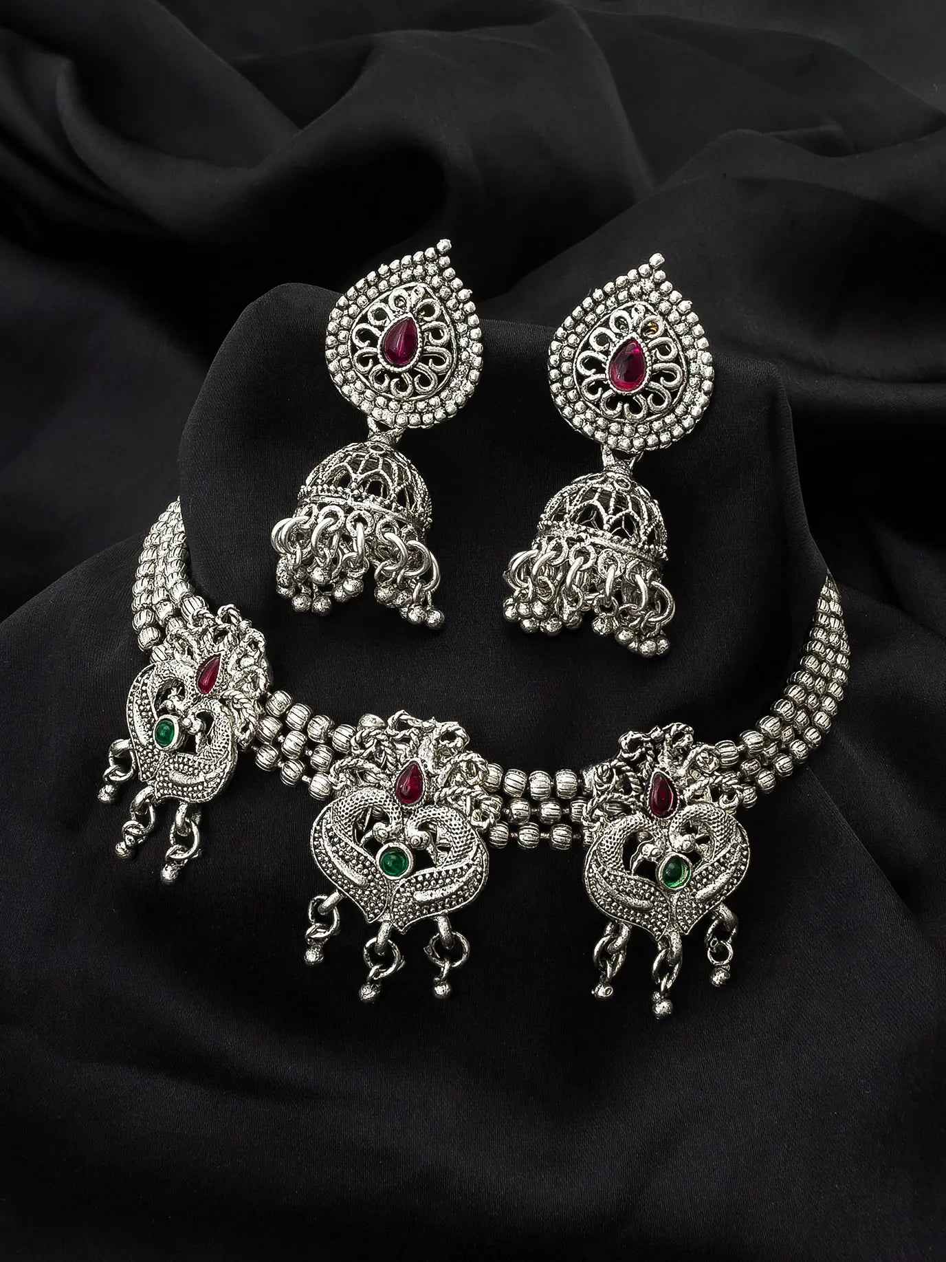 Silver-toned Indian jewelry set featuring a choker necklace, matching earrings, and a headpiece, decorated with intricate filigree work and small red and green gemstones, displayed on a black fabric background.