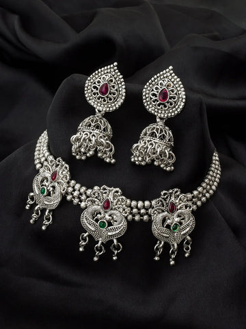 Silver-toned Indian jewelry set featuring a choker necklace, matching earrings, and a headpiece, decorated with intricate filigree work and small red and green gemstones, displayed on a black fabric background.