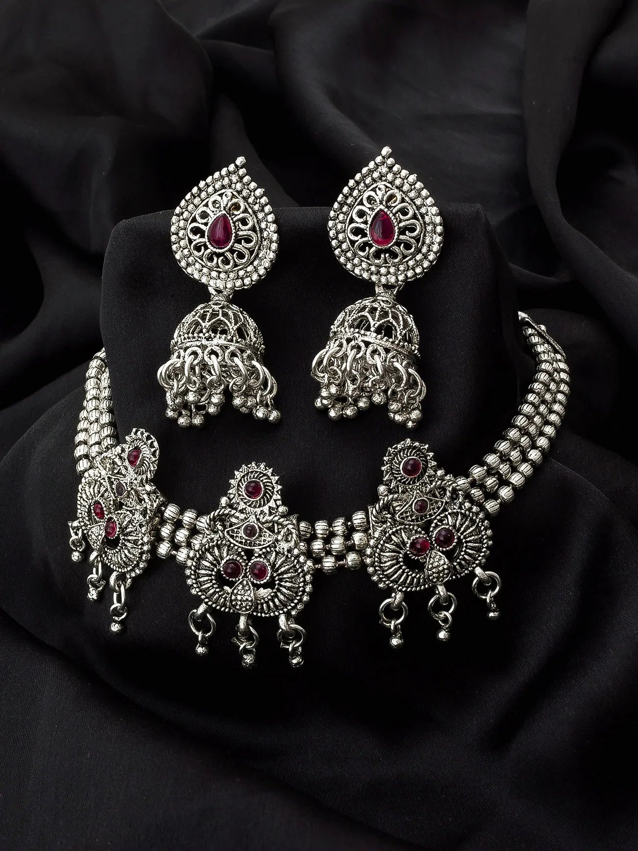 Silver-toned Indian jewelry set featuring a choker necklace, matching earrings, and a bangle, decorated with intricate filigree work and small red gemstone accents, displayed on a black fabric background.