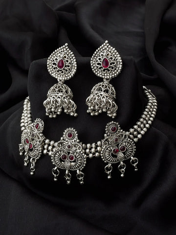 Silver-toned Indian jewelry set featuring a choker necklace, matching earrings, and a bangle, decorated with intricate filigree work and small red gemstone accents, displayed on a black fabric background.