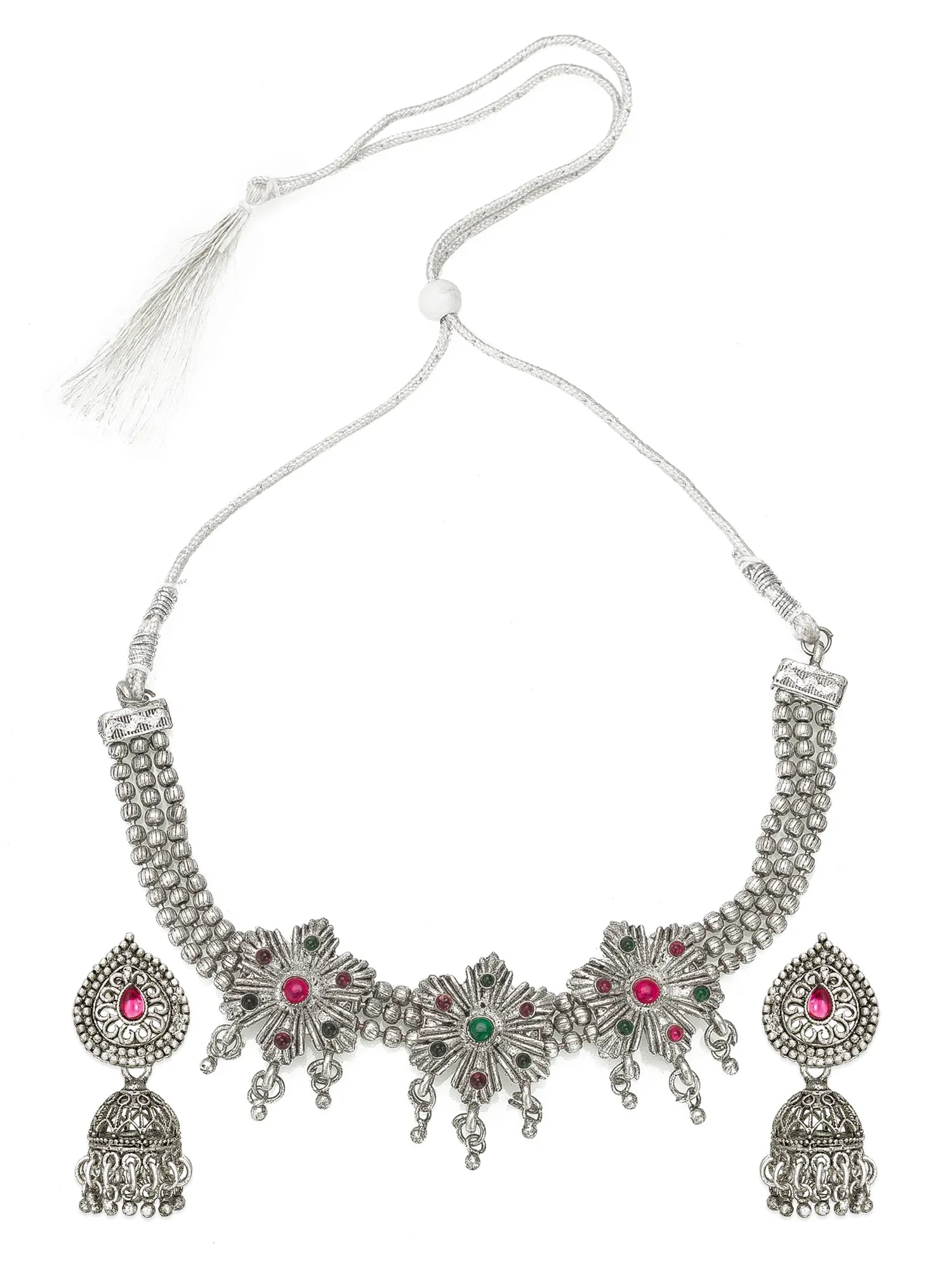 An ornate silver-toned traditional necklace with matching jhumka earrings, featuring silver floral motifs set with pink and green stones, suspended from a silver cord with a tassel, on a plain white background.