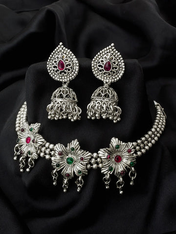 An ornate silver-toned jewelry set displayed on a black fabric background, including a choker necklace and matching earrings. The pieces feature intricate filigree detailing, beaded borders, and red and green stone accents.