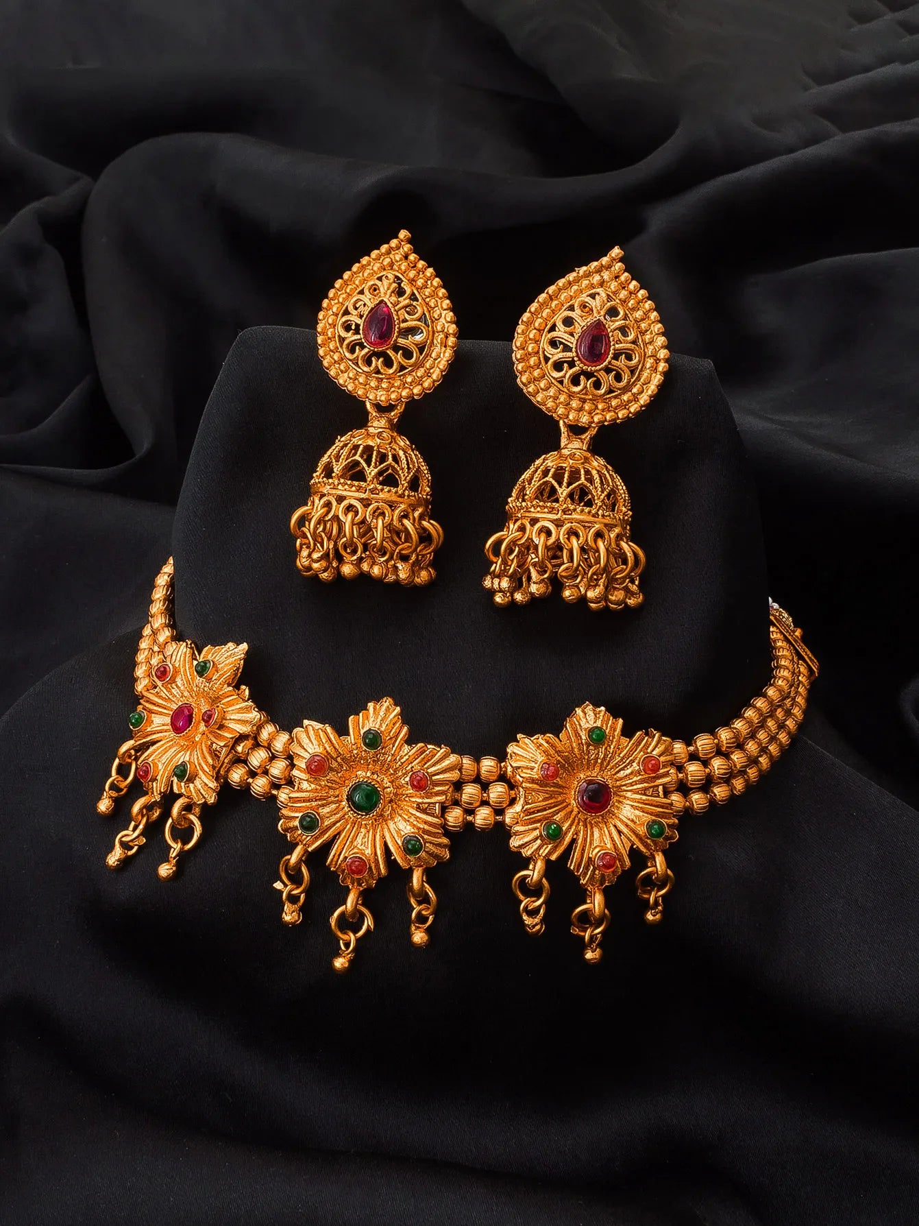 Gold-toned traditional Indian jewelry set featuring a choker necklace, matching earrings, and a small pendant, decorated with floral motifs and red and green stone accents, displayed on a black fabric background.