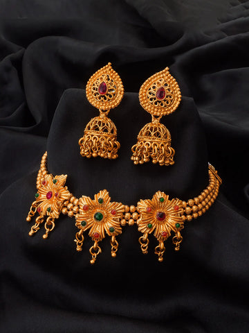Gold-toned traditional Indian jewelry set featuring a choker necklace, matching earrings, and a small pendant, decorated with floral motifs and red and green stone accents, displayed on a black fabric background.