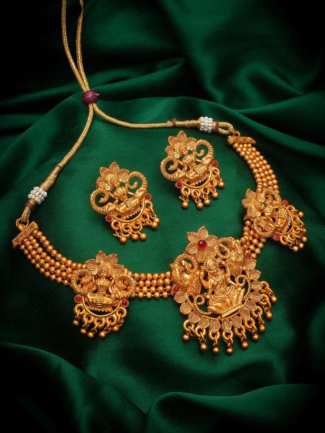 An ornate gold-toned traditional jewelry set displayed on draped dark green satin fabric, including a necklace, earrings, and matching pendant pieces with floral and filigree details.