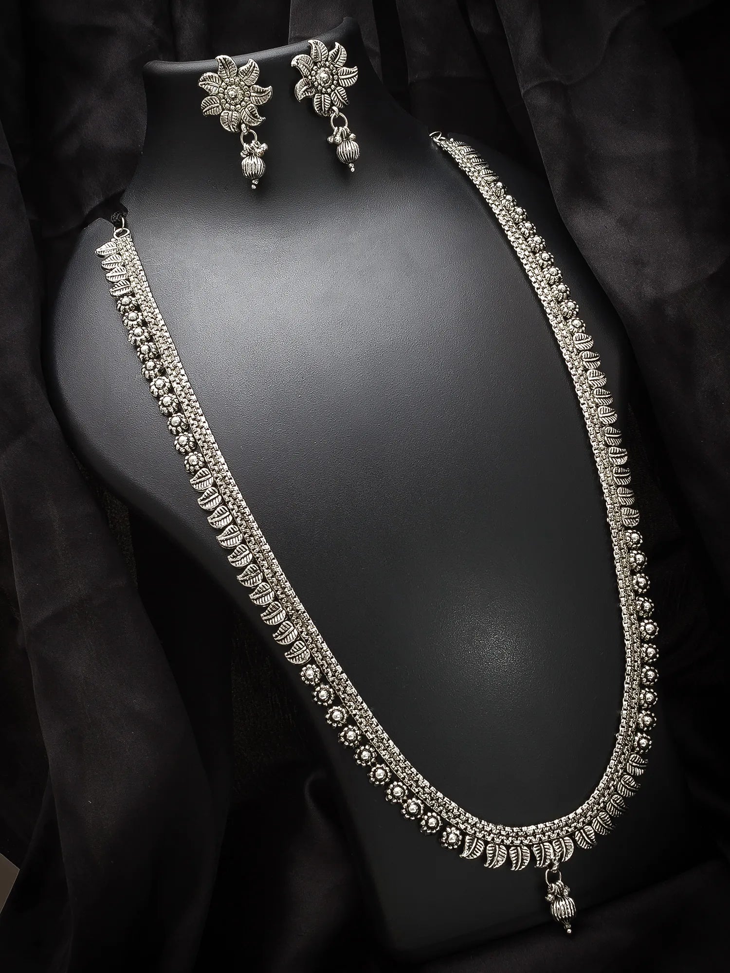A silver-toned traditional Indian jewelry set featuring a necklace, matching earrings, and a bracelet displayed on a black mannequin bust against a black fabric backdrop.