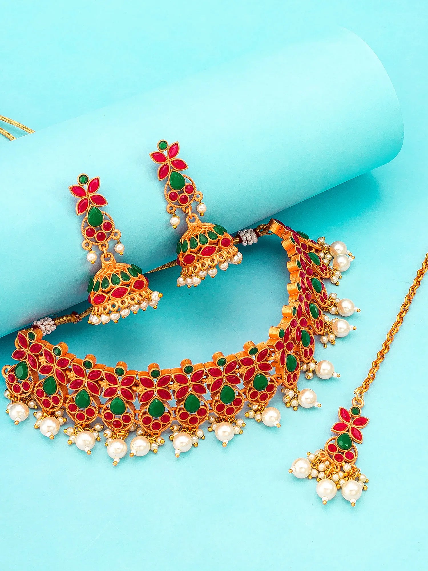 Gold-plated Indian bridal jewelry set featuring a choker necklace, matching jhumka earrings, and a maang tikka, decorated with red and green enamel, white faux pearls, and set against a light blue background.