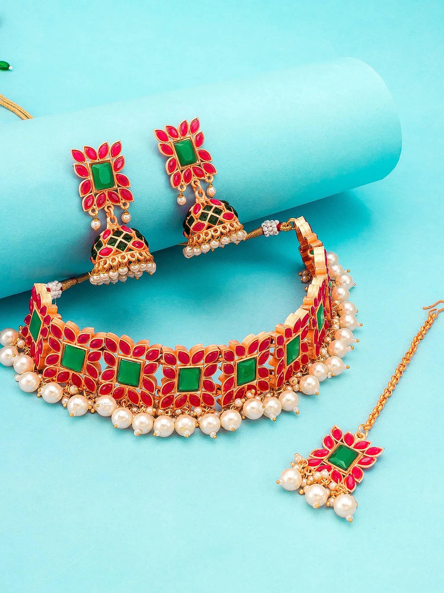 Gold-plated Indian jewelry set featuring a choker necklace, matching earrings, and a maang tikka, decorated with red floral inlays, green square stones, and white faux pearls, displayed on a light blue background.