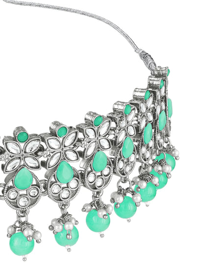An ornate silver-toned statement necklace featuring intricate floral filigree, small clear stones, and dangling green teardrop and round beads on a white background.