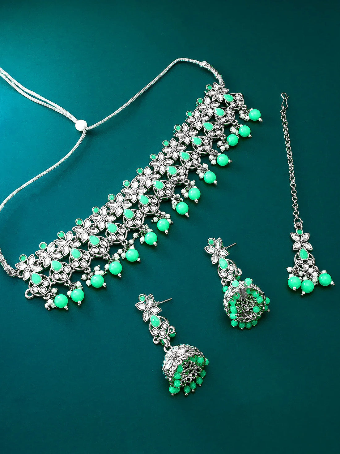 Silver-toned jewelry set with green stones, including a necklace, matching earrings, and a chain, arranged on a dark teal background.