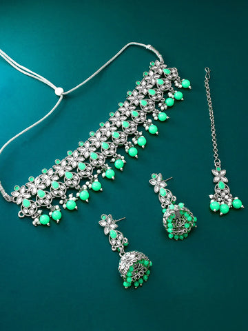 Silver-toned jewelry set with green stones, including a necklace, matching earrings, and a chain, arranged on a dark teal background.